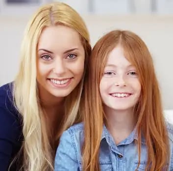 Lächelnde blonde Mutter und rothaarige Tochter sitzen nah beieinander.