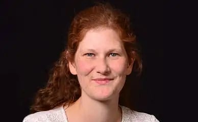 Porträt einer Lehrerin mit lockigem roten Haar und weißem Oberteil vor schwarzem Hintergrund.