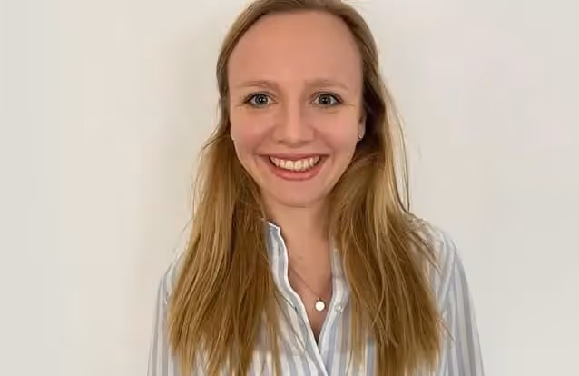 Lehrerin mit langem blonden Haar, die lächelnd vor einem weißen Hintergrund steht und ein gestreiftes Hemd trägt.