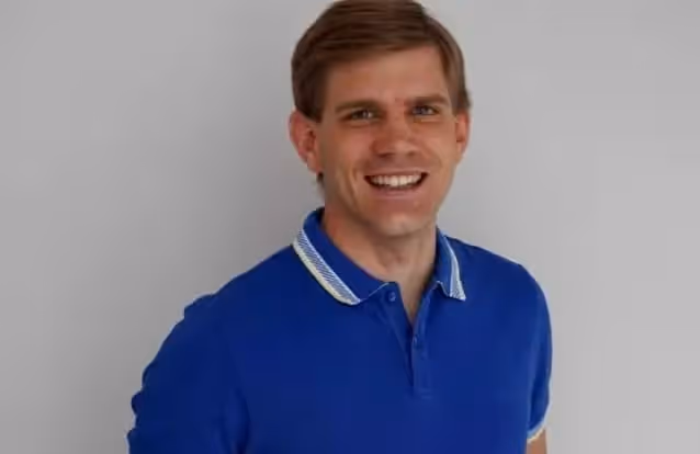 Lächelnder Lehrer mit kurzem braunem Haar trägt ein blaues Poloshirt vor grauem Hintergrund.