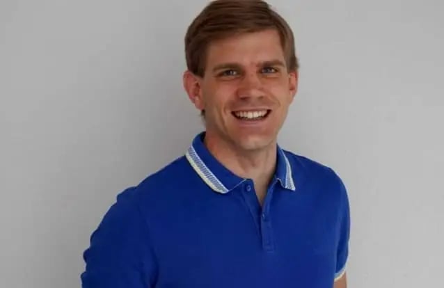 Lächelnder Lehrer mit kurzem braunem Haar trägt ein blaues Poloshirt vor grauem Hintergrund.