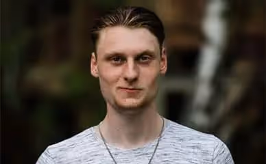 Junger Lehrer mit hellbraunen Haaren trägt ein hellgraues T-Shirt und steht vor unscharfem Waldhintergrund.