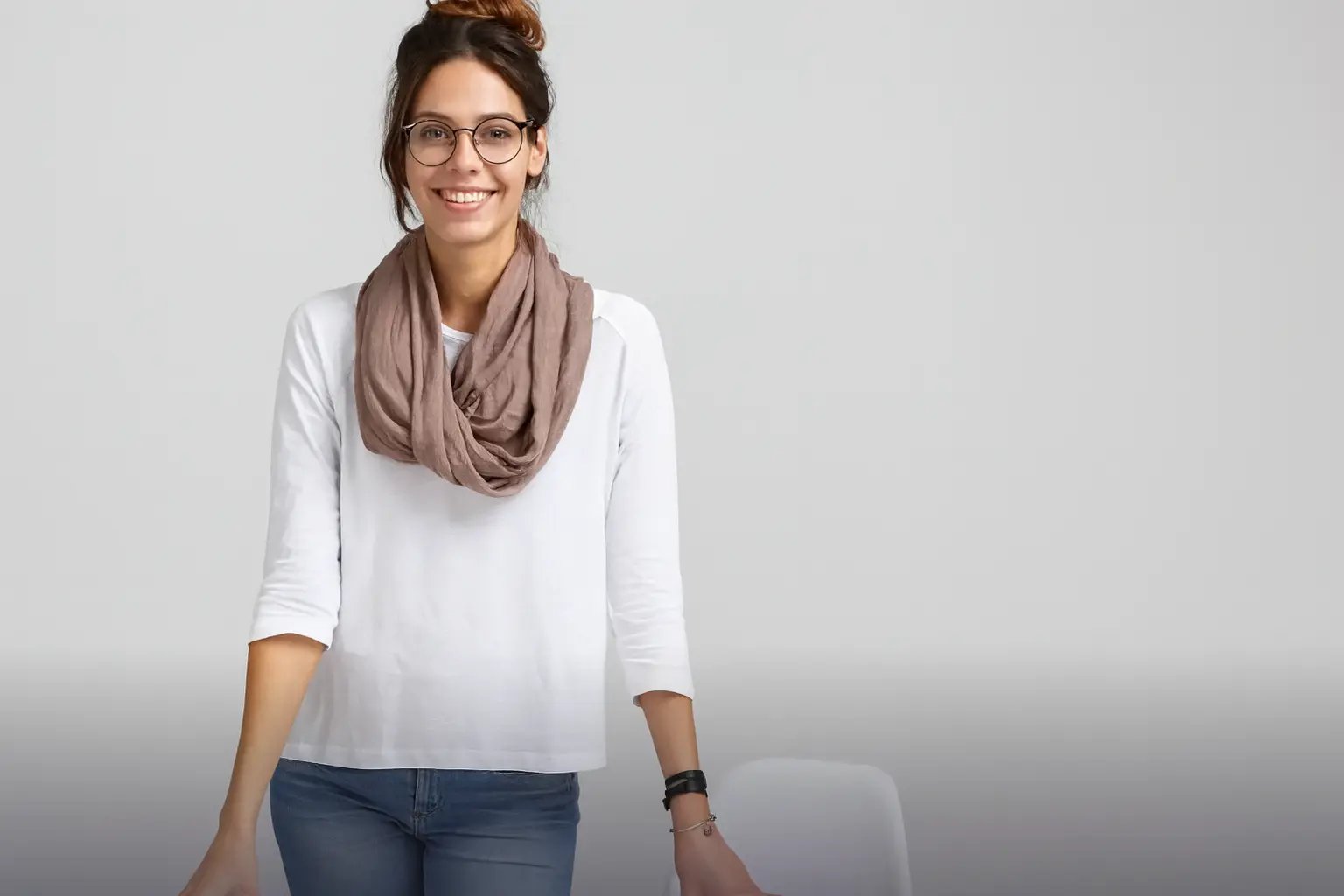Junge Frau mit Brille, weißem Oberteil, braunem Schal und Hochsteckfrisur lächelt vor grauem Hintergrund.