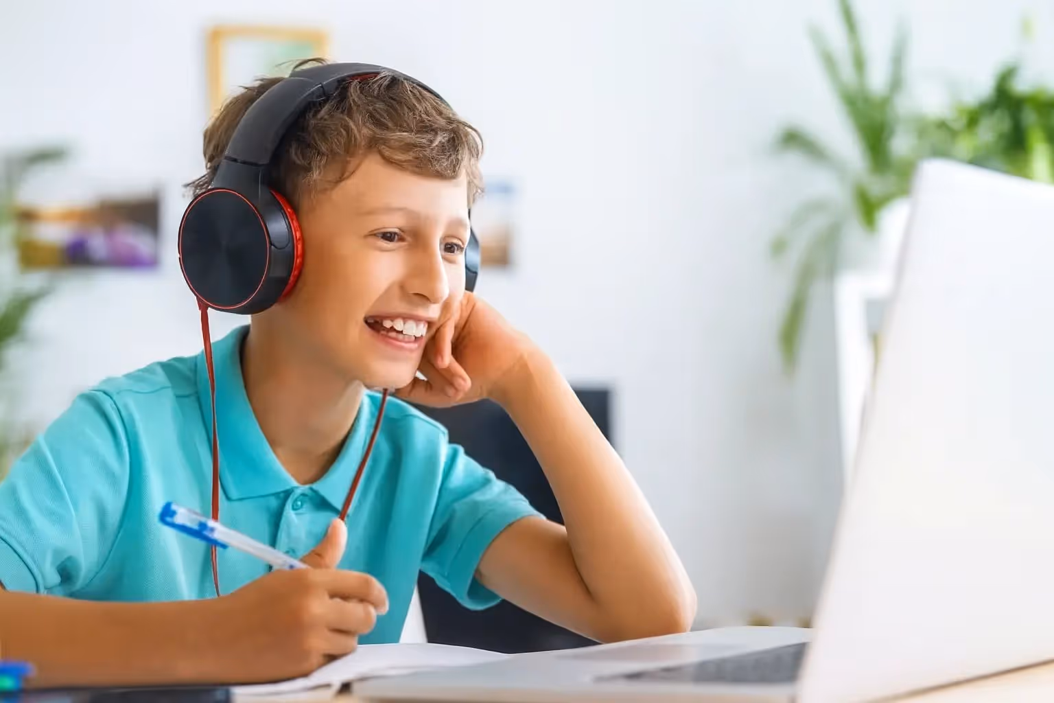 Schüler mit Kopfhörern und Stift sitzt lächelnd vor einem Laptop und macht Hausaufgaben.