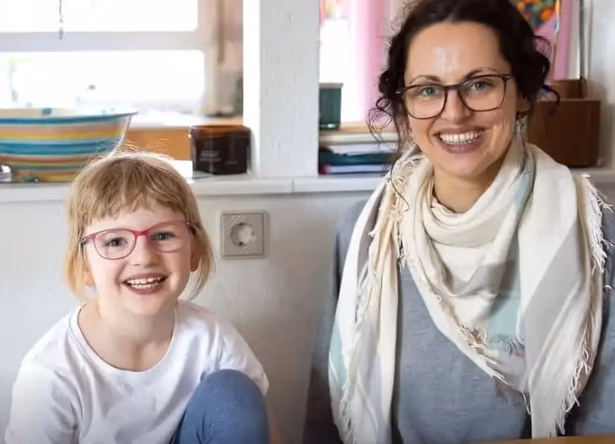 Eine lächelnde Mutter mit Brille und Schal sitzt neben einem lachenden Mädchen mit roter Brille und weißem T-Shirt im Wohnzimmer.