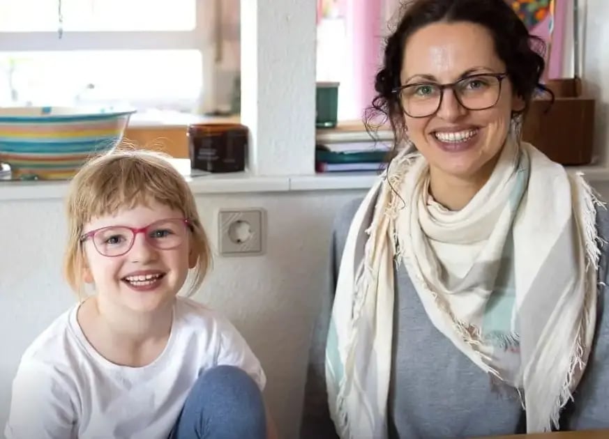 Eine lächelnde Mutter mit Brille und Schal sitzt neben einem lachenden Mädchen mit roter Brille und weißem T-Shirt im Wohnzimmer.