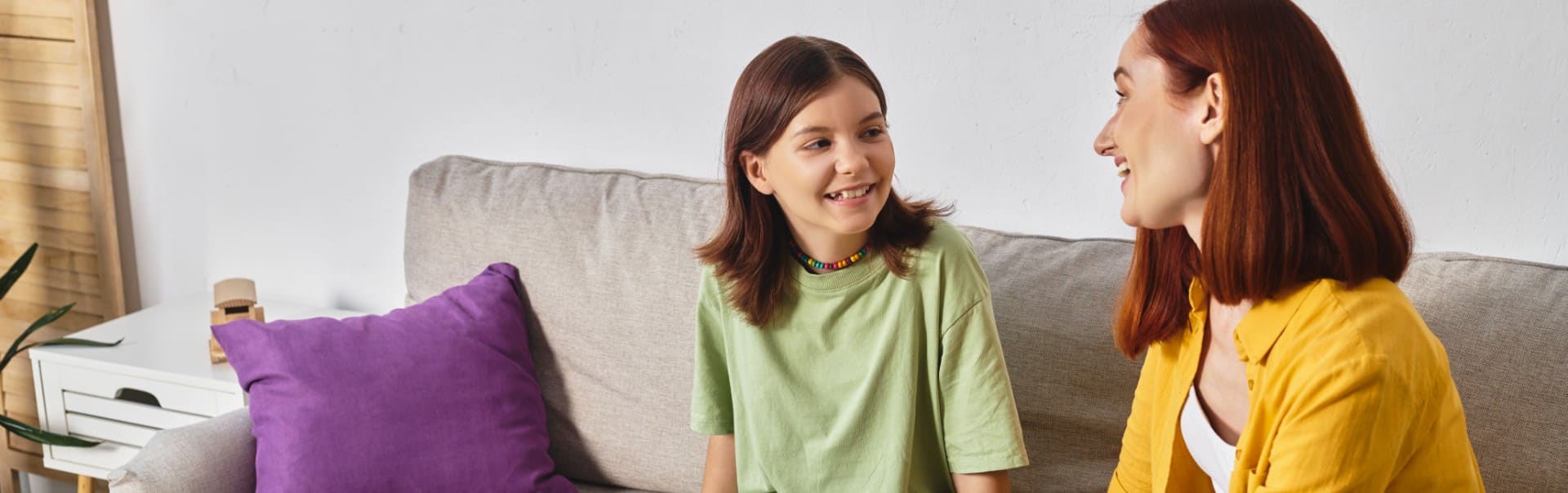 Zwei Frauen, eine mit braunen Haaren und grünem T-Shirt, die andere mit roten Haaren und gelbem Hemd, sitzen auf einem Sofa und unterhalten sich.