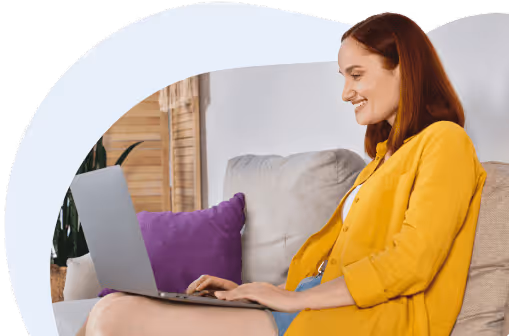 Mutter in gelbem Hemd, die auf einem beigen Sofa sitzt und auf einem Laptop arbeitet.