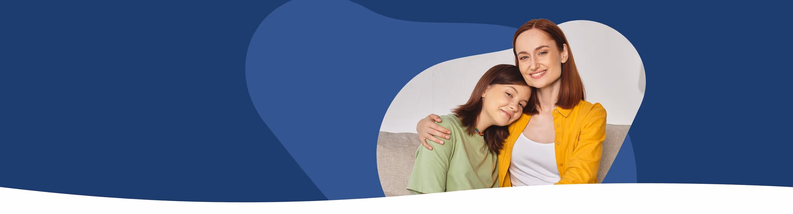 Eine lächelnde Mutter mit rotem Haar in gelber Jacke umarmt ihre Tochter in grüner Kleidung auf einem Sofa.