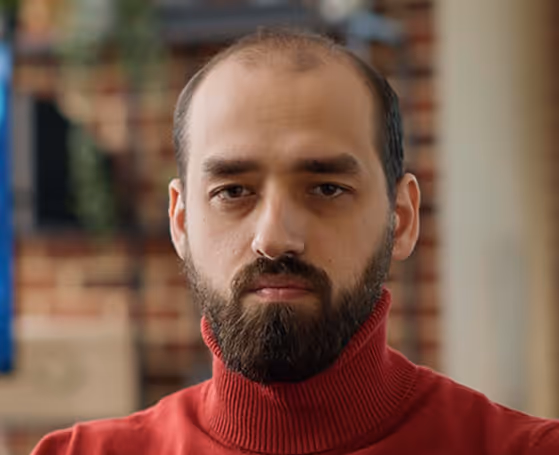 Mann mit Bart und zurückgehendem Haar trägt roten Rollkragenpullover vor unscharfem Hintergrund.