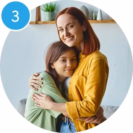 Mutter in gelbem Hemd umarmt lächelnd ihre Tochter in grünem T-Shirt in einem hellen Wohnzimmer.