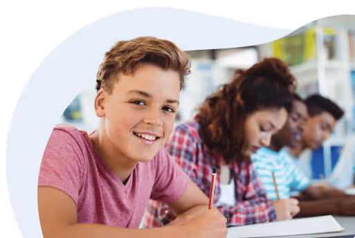 Lächelnder Schüler in einem rosa T-Shirt schreibt in einem Klassenzimmer, während andere Schüler im Hintergrund konzentriert arbeiten.