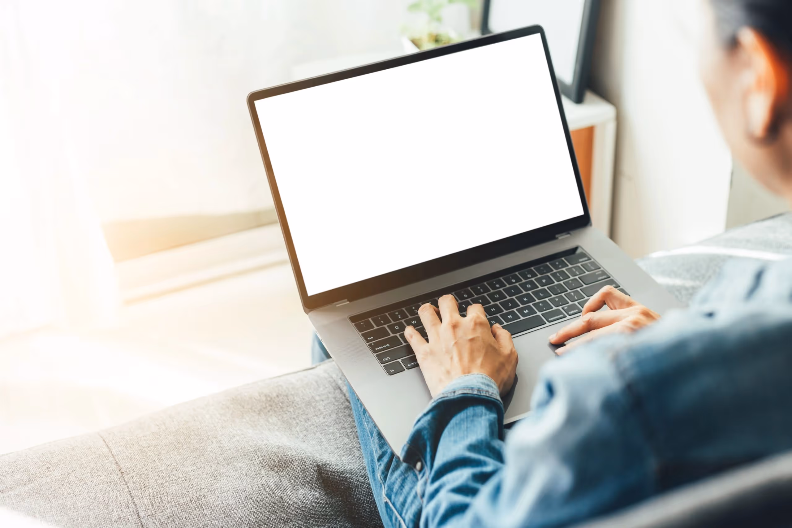 Person tippt auf einem Laptop mit leerem Bildschirm, sitzend auf einem grauen Sofa in hellem Raum.