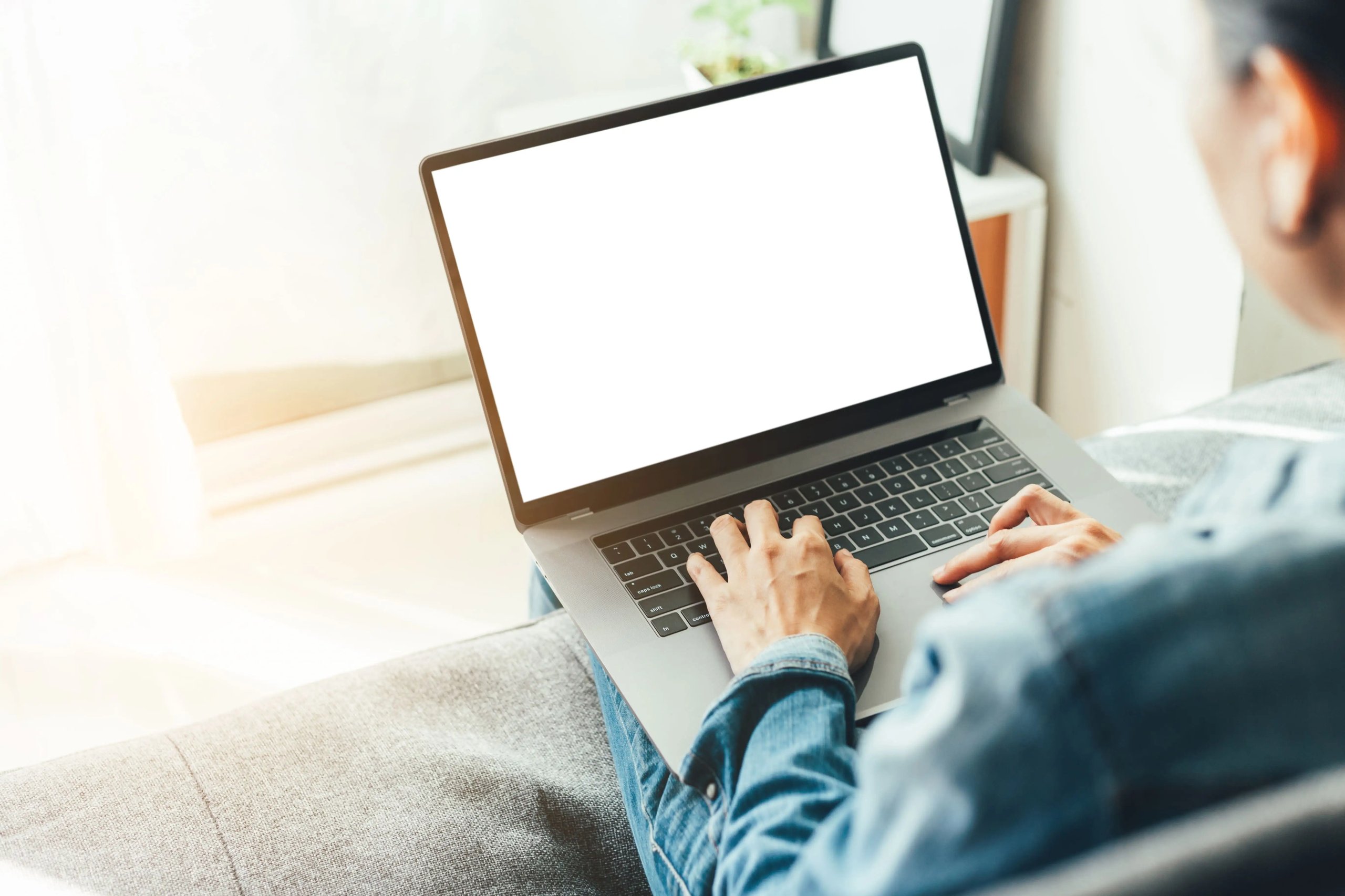 Person tippt auf einem Laptop mit leerem Bildschirm, sitzend auf einem grauen Sofa in hellem Raum.