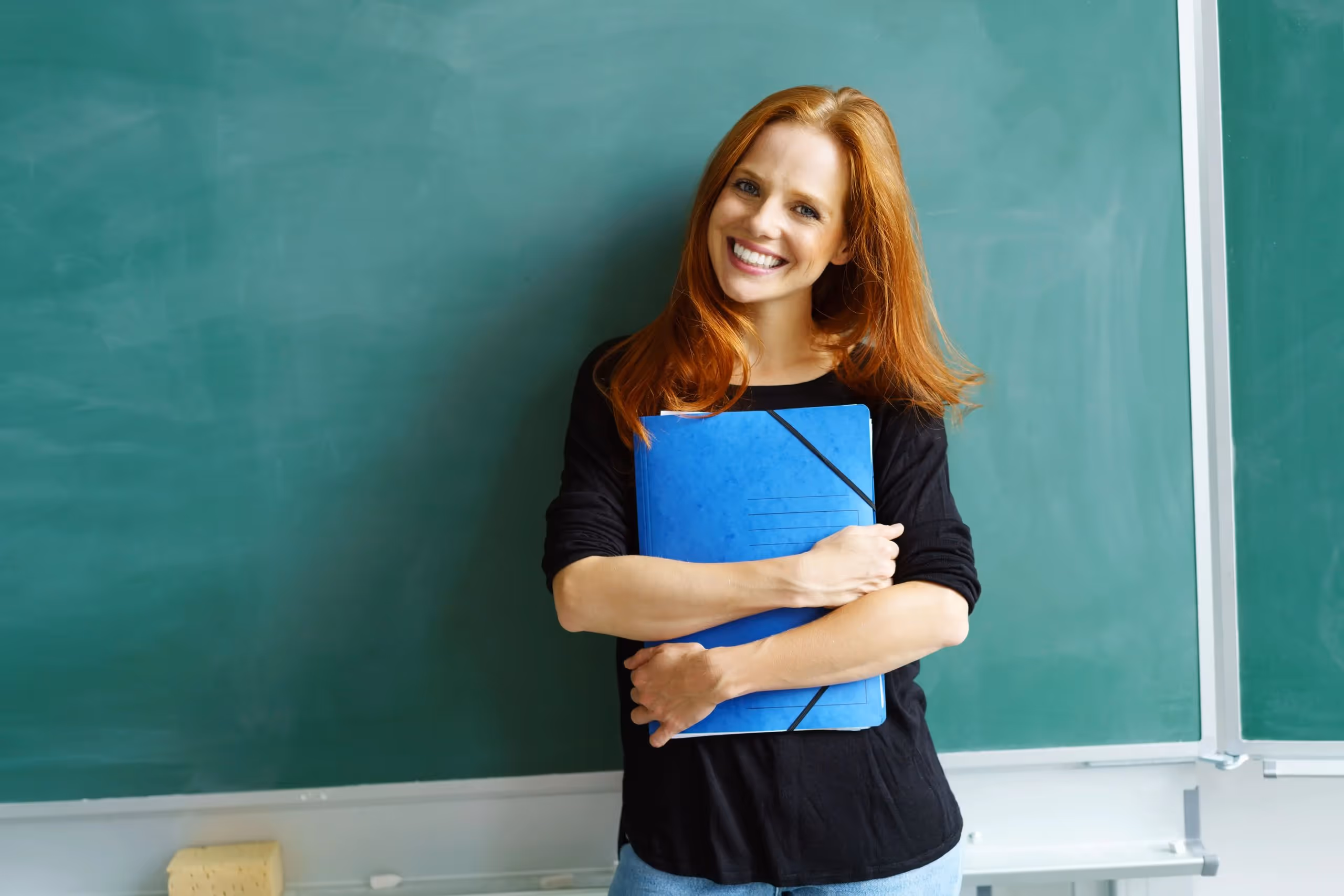 Lächelnde Frau mit roten Haaren hält blauen Ordner vor einer grünen Schultafel.