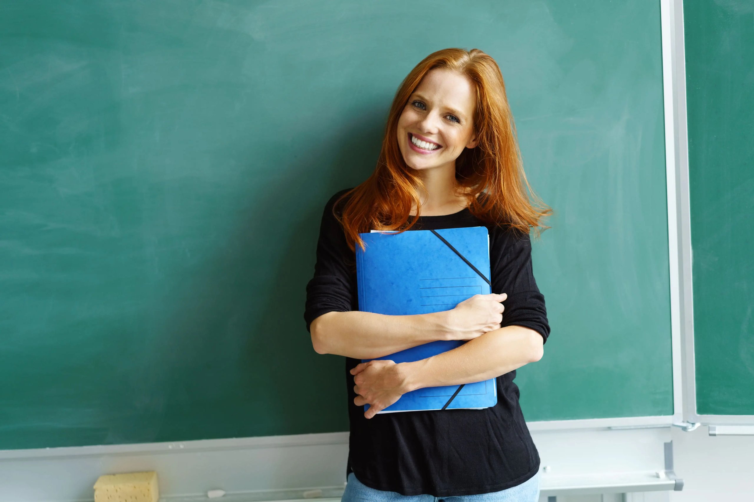Lächelnde Frau mit roten Haaren hält blauen Ordner vor einer grünen Schultafel.