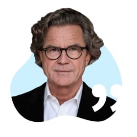 Älterer Mann mit lockigem Haar, Brille und Anzug vor blauem Hintergrund mit Anführungszeichen.