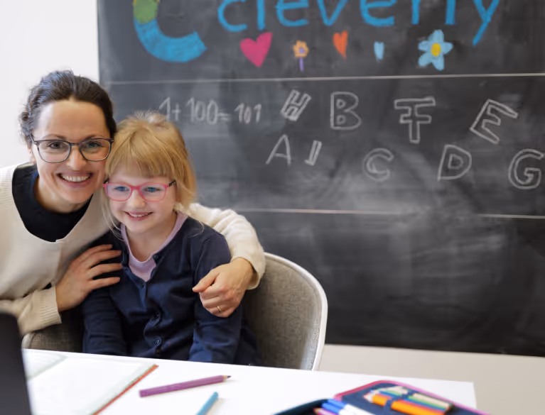 Frau umarmt lachendes Mädchen mit rosaroter Brille vor einer Schultafel mit Buchstaben und einer Rechenaufgabe.