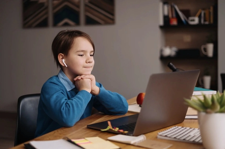 Ein Junge mit blauer Strickjacke sitzt an einem Schreibtisch, schaut auf einen Laptop und trägt kabellose Kopfhörer.