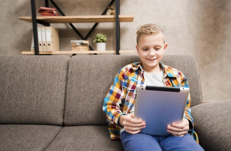Ein lächelnder Junge sitzt auf einem grauen Sofa und hält ein Tablet in den Händen.