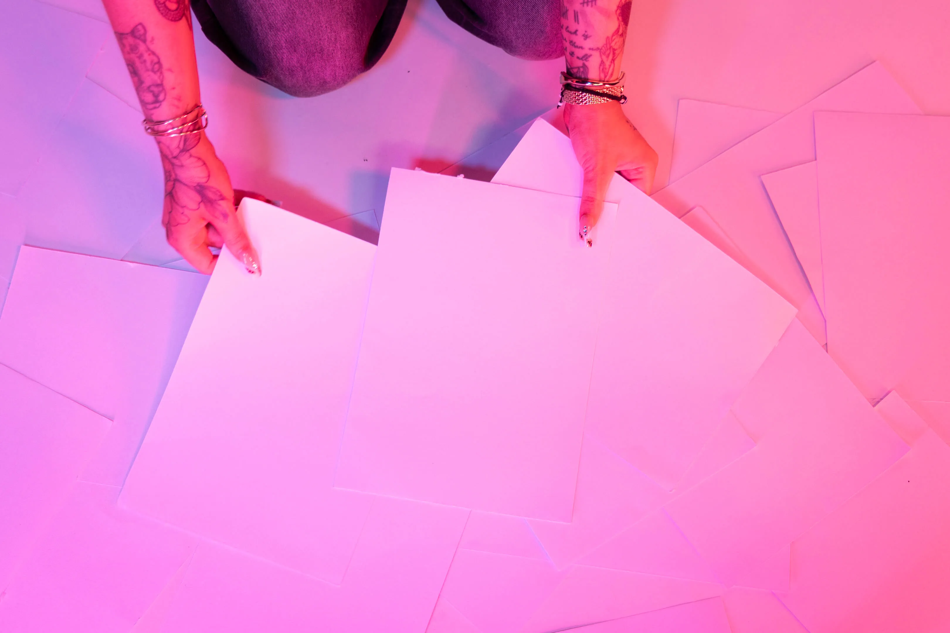 Two tattooed hands with bracelets holding blank white sheets of paper over numerous scattered sheets, under pink and purple lighting.