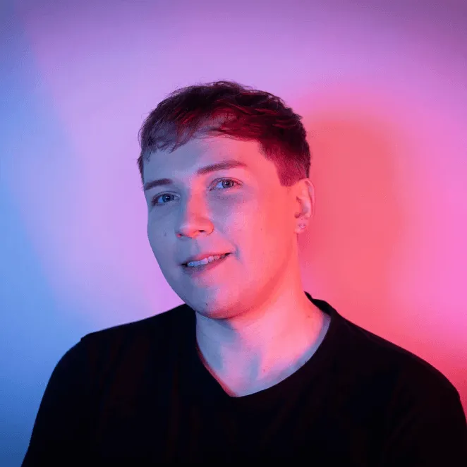 Portrait of a young person with short hair, wearing a black shirt, illuminated by blue and pink lighting against a pink-purple background.