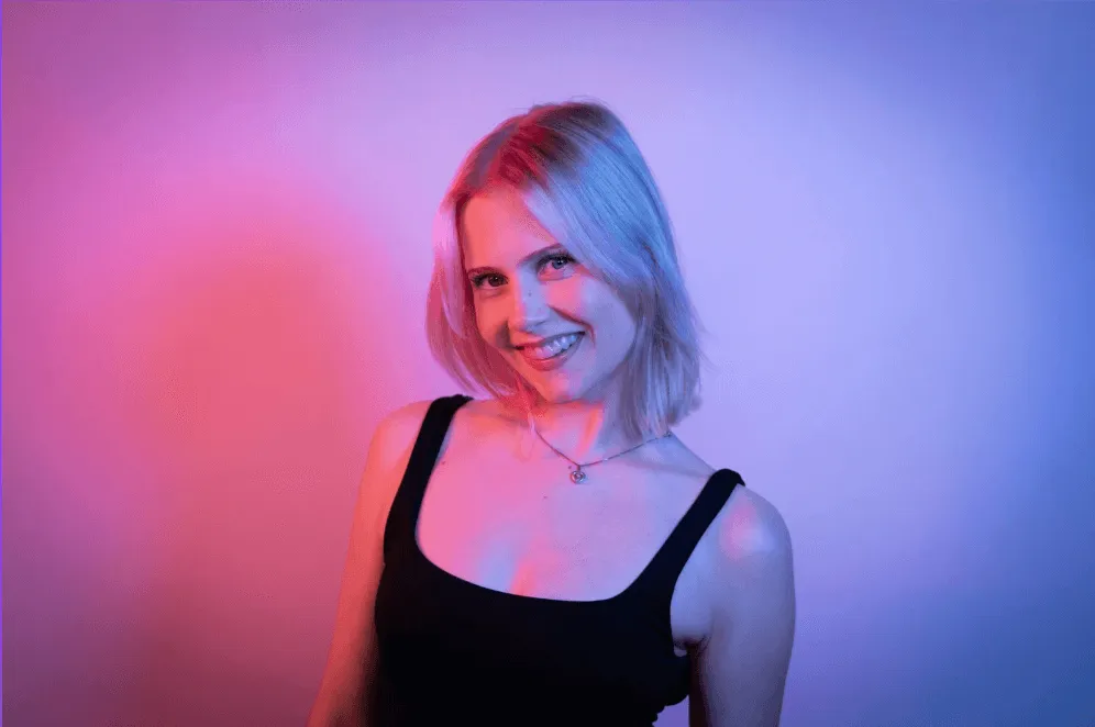 Smiling woman with short blonde hair wearing a black tank top, illuminated by pink and blue lighting against a plain background.