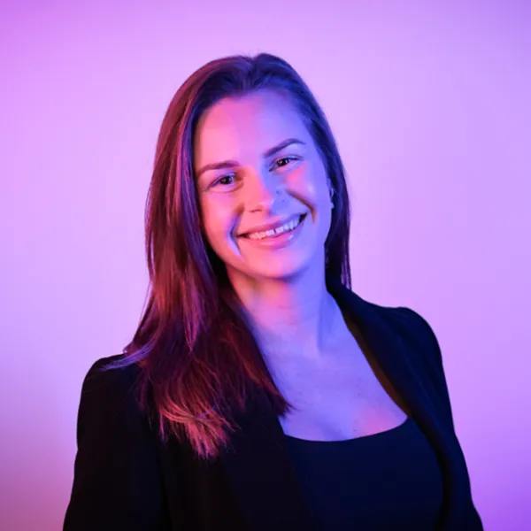 Smiling woman with long, wavy hair illuminated by pink and blue lighting, wearing a black top.