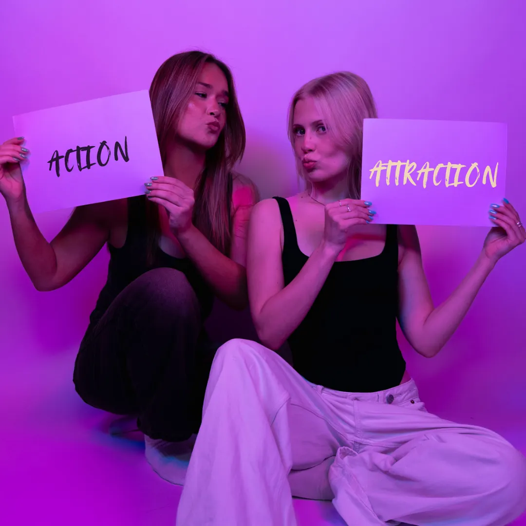 Two women posing and holding signs that say 'ACTION' and 'ATTRACTION' under purple lighting.