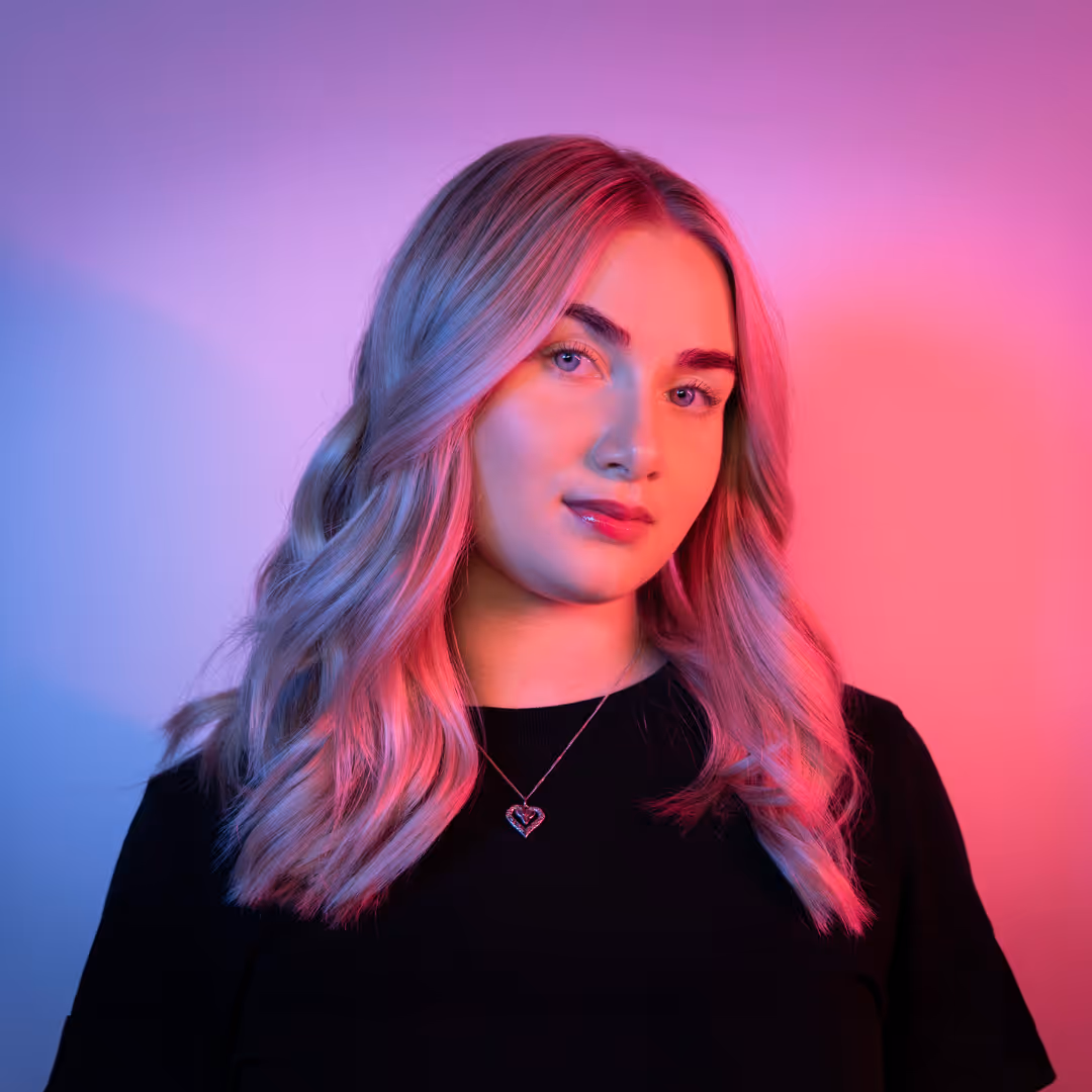 Smiling woman with long, wavy hair illuminated by pink and blue lighting, wearing a black top.