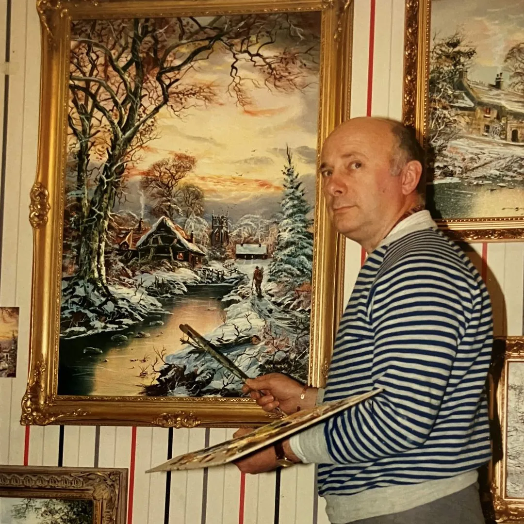John Corcoran holding a paint palette and brush, standing next to a framed winter landscape painting with a snowy village and stream.