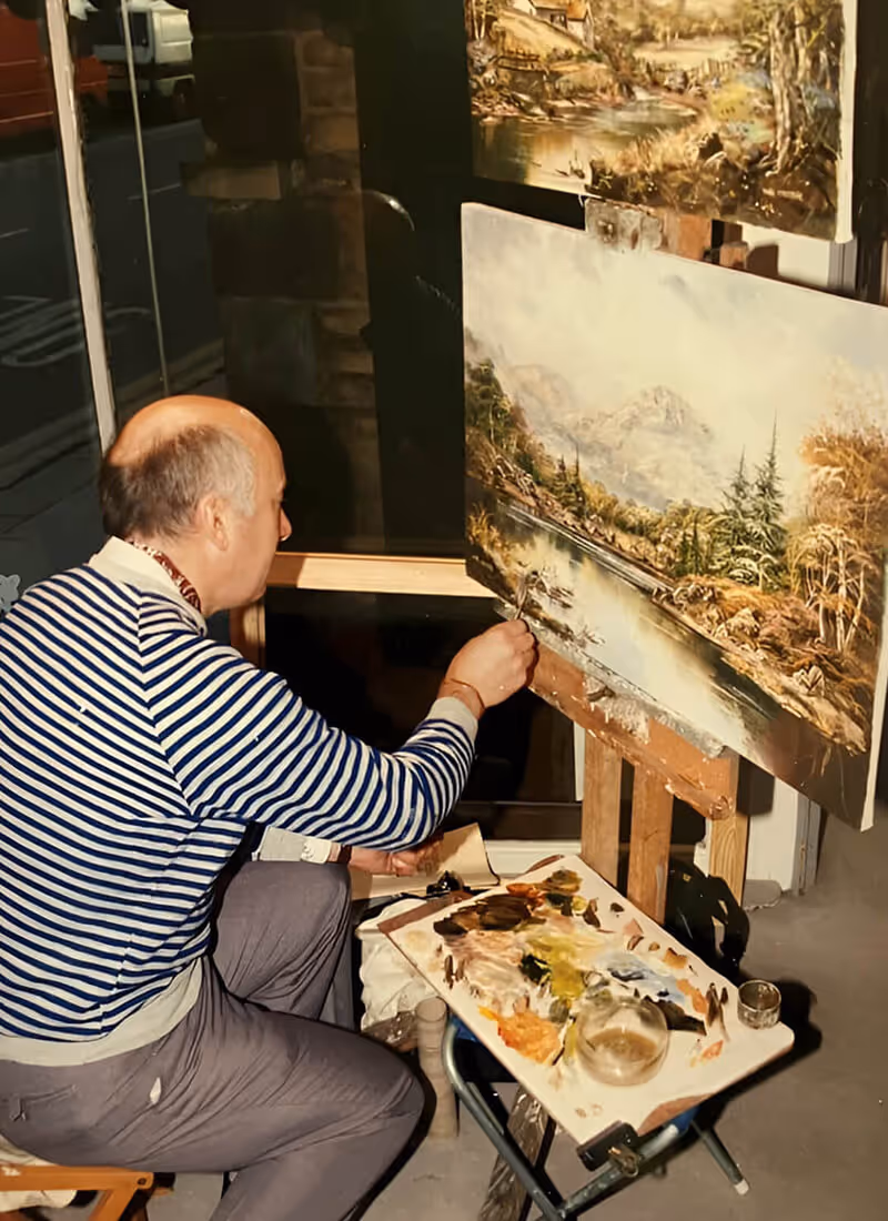 John Corcoran painting a detailed landscape of trees, river, and mountains on a canvas set on an easel.