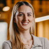 Smiling woman with long blonde hair wearing a light gray top and a necklace in a warmly lit interior.