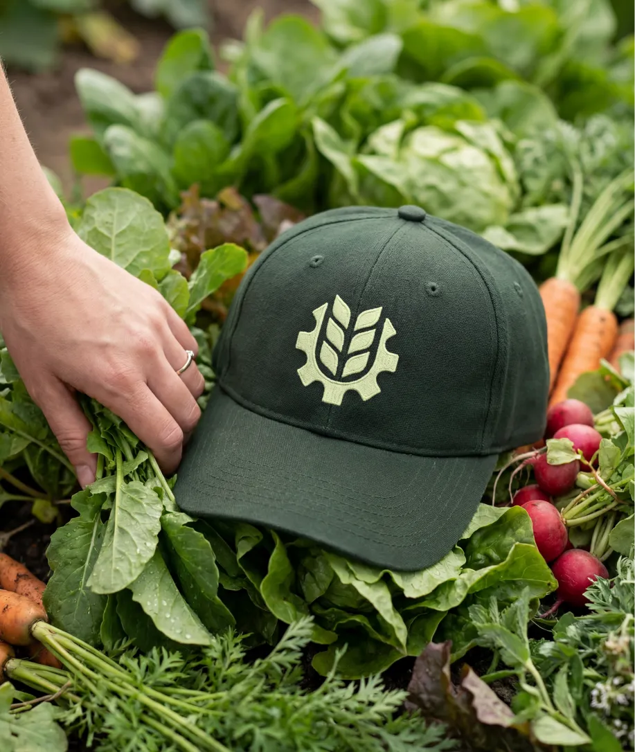 Green baseball cap with a white gear and leaf logo resting on fresh leafy greens surrounded by carrots and radishes, with a hand picking leaves.