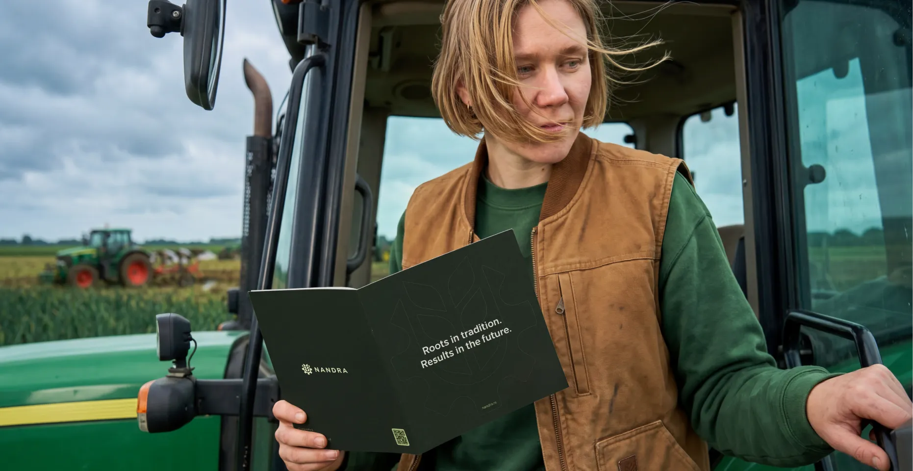 Person in green shirt and brown vest holding an open brochure that reads 'Roots in tradition. Results in the future.' standing beside a green tractor in a field.