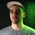 Young man wearing a light-colored cap and gray shirt, illuminated by green and neutral lighting against a dark background.