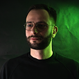 Portrait of a bearded man with short hair, wearing a green jacket and black shirt, with green and black background lighting.
