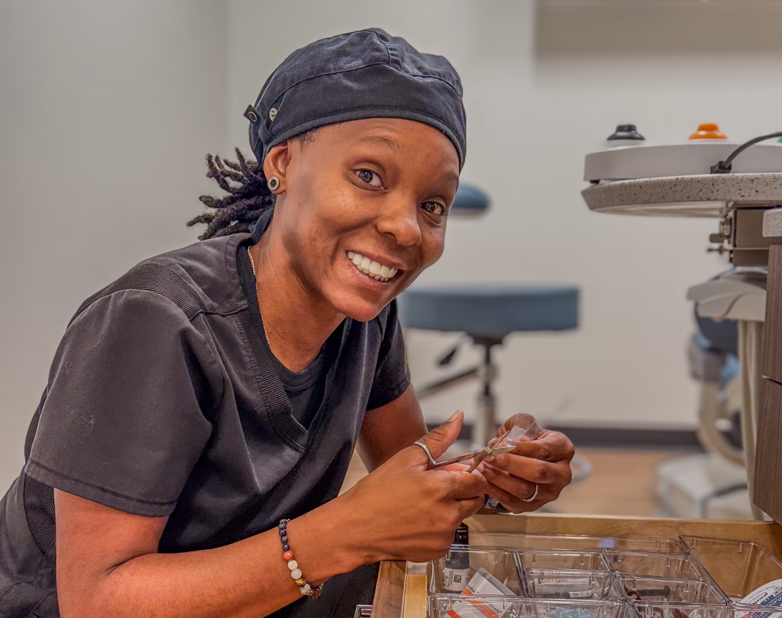 Smiling dental professional in scrubs holding instruments beside a sterilization tray.