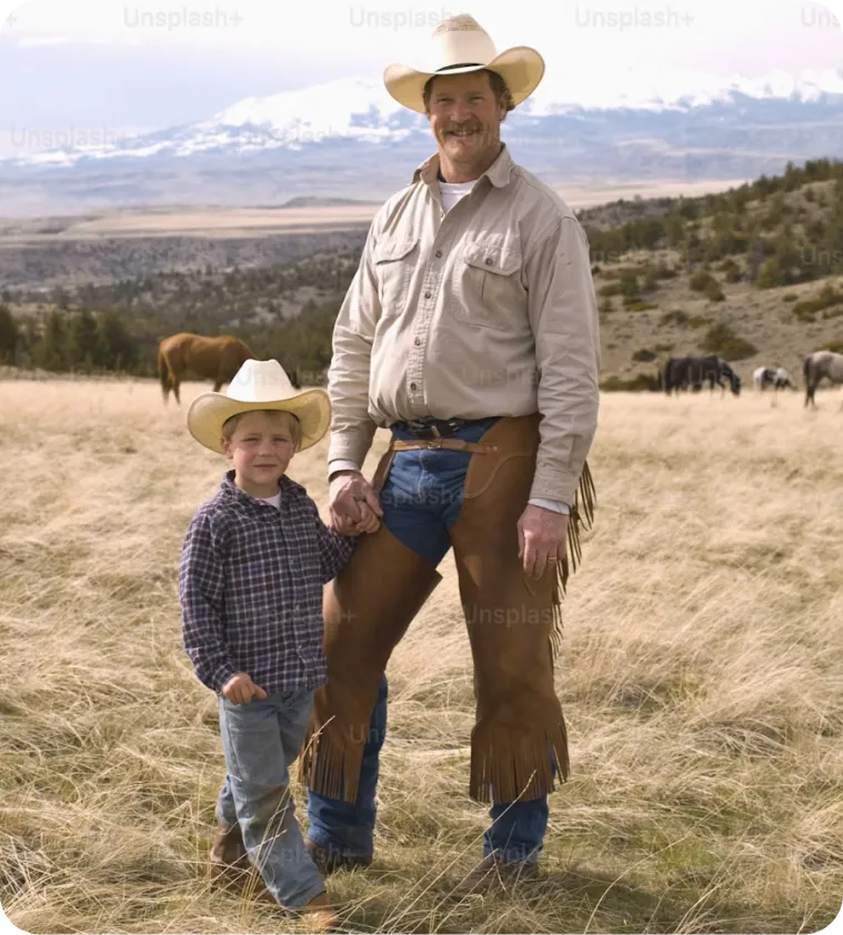 Cowboy standing with a young child in an open pasture with grazing cattle and mountains in the distance
