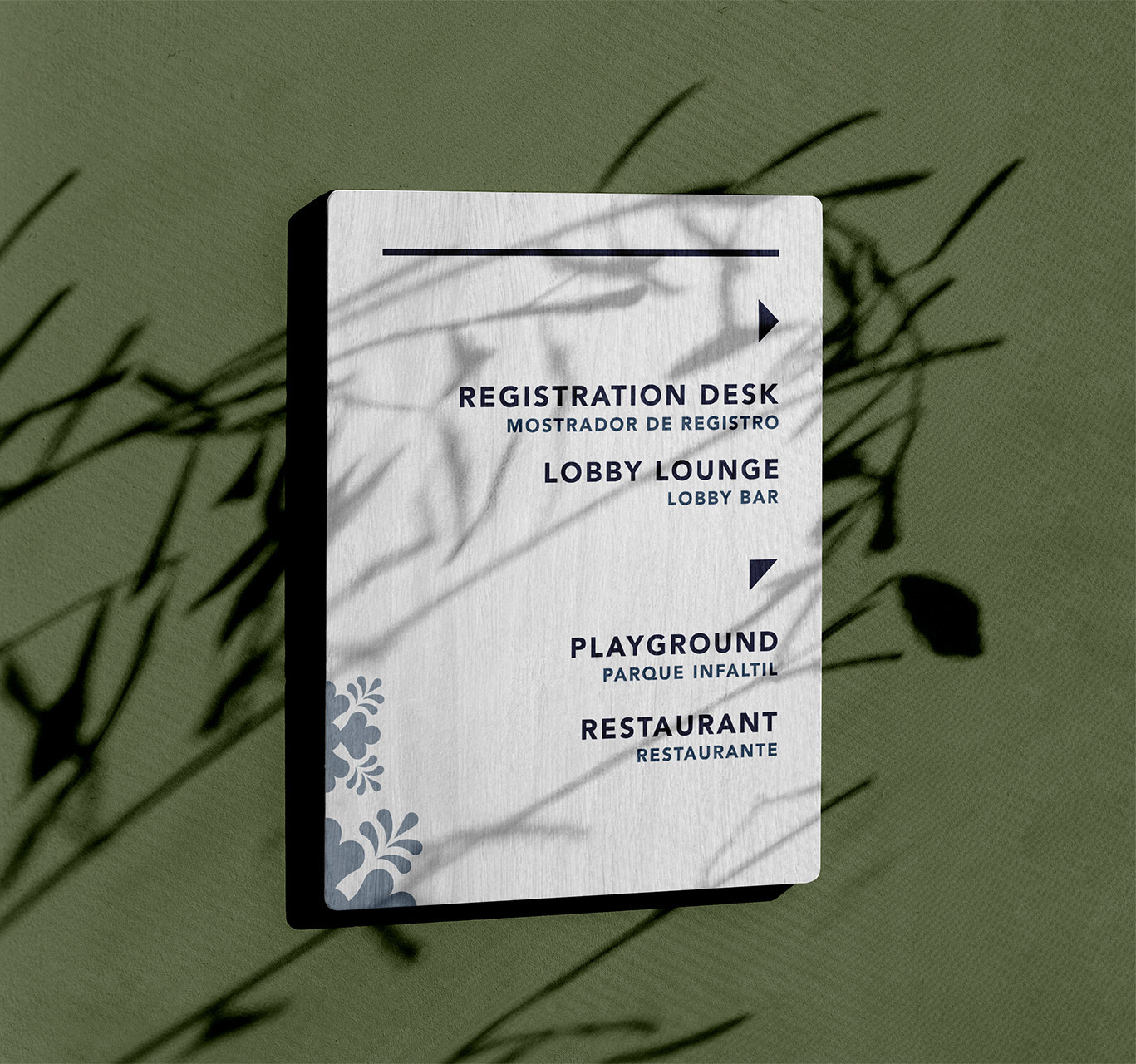 White directional sign with black text and arrows on green background, indicating the registration desk, lobby lounge, playground, and restaurant with Spanish translations.