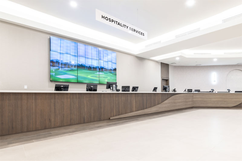 Modern hospitality services reception area with long wooden desk, multiple computer monitors, and large wall screen displaying a golf course.