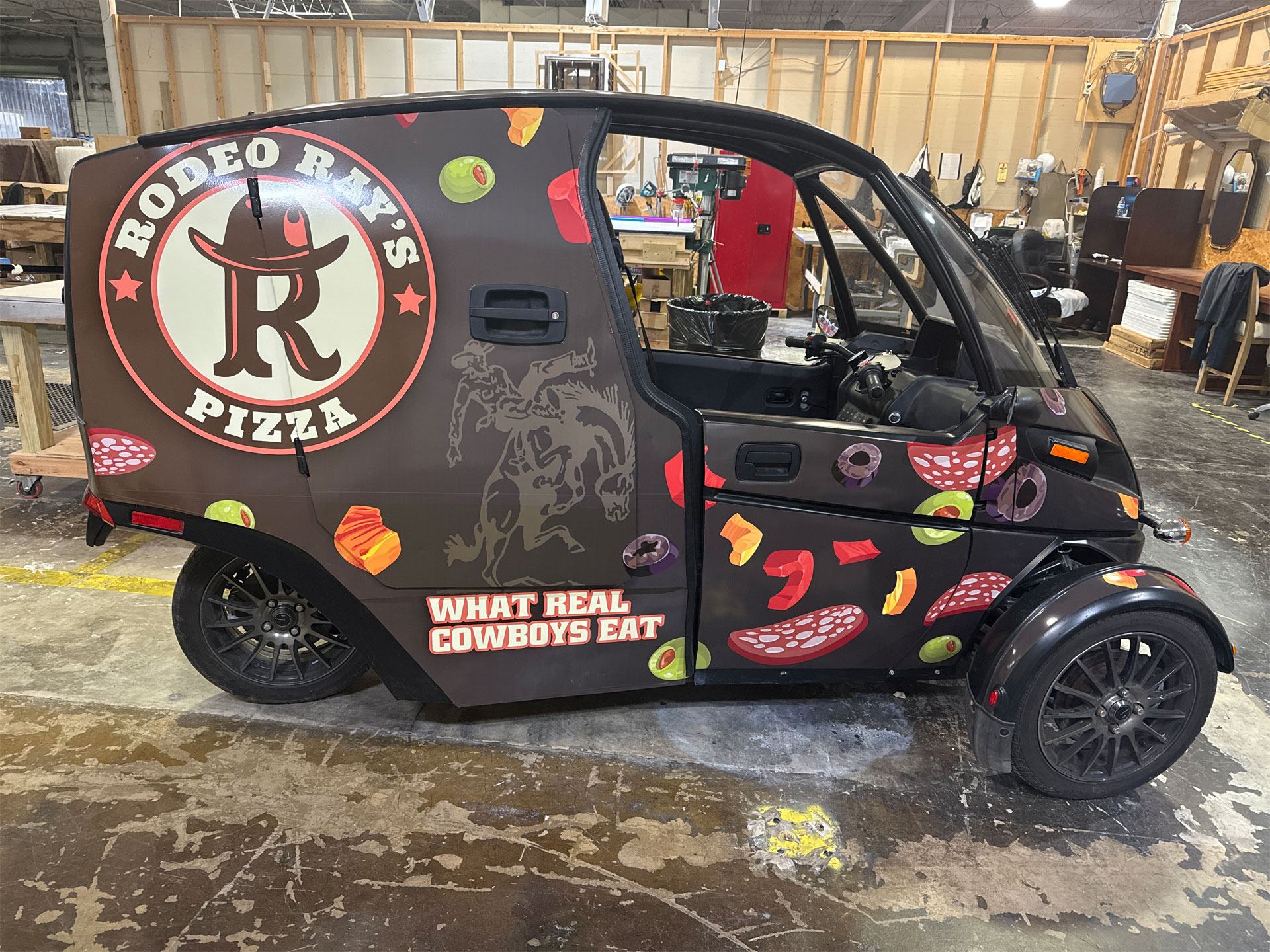 Small black vehicle with Rodeo Ray's Pizza logo, pizza toppings graphics, and slogan 'What Real Cowboys Eat' inside a workshop.