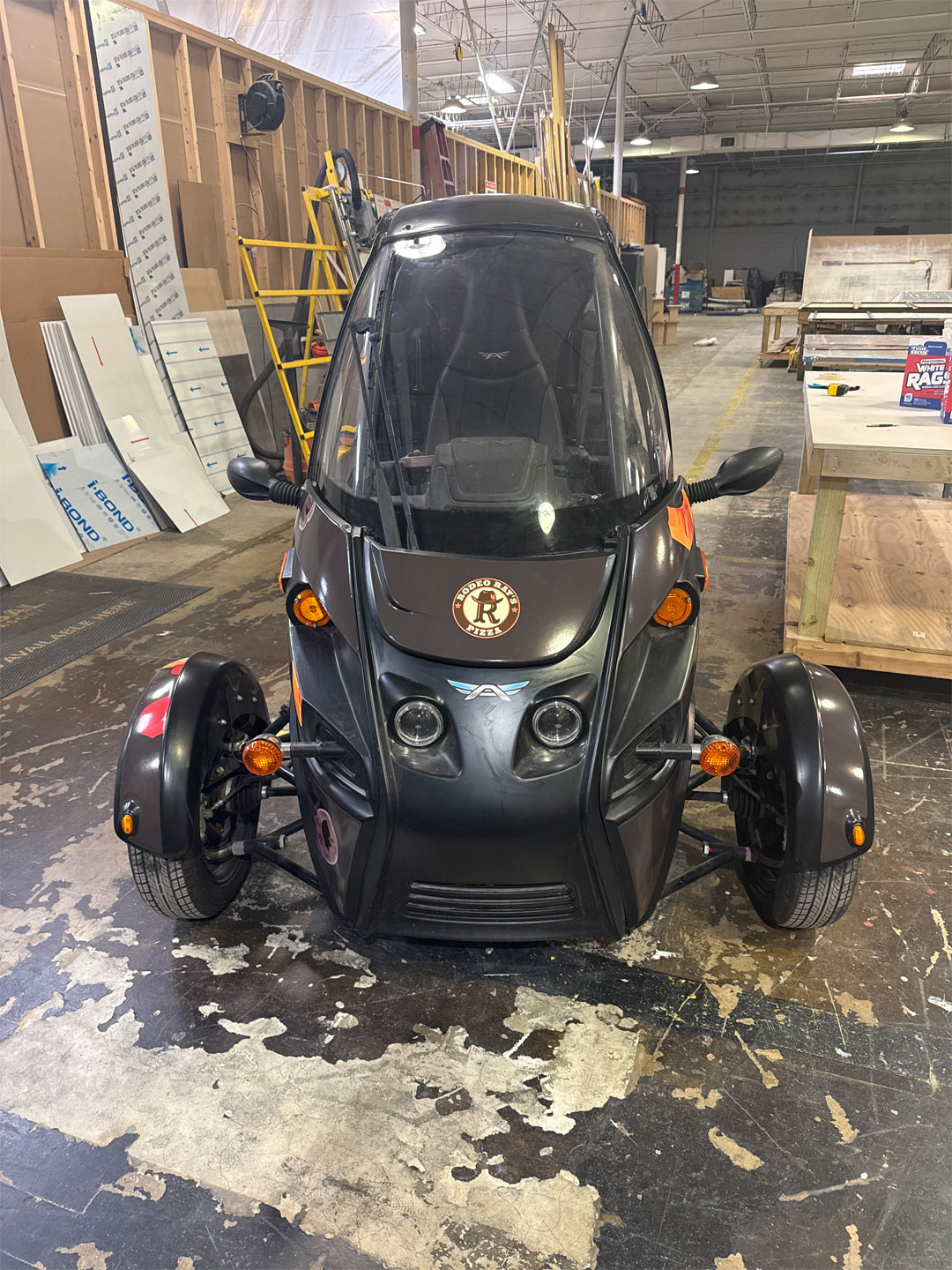 Front view of a small black three-wheeled enclosed vehicle with 'Rodeo Ray's Pizza' logo on the hood inside a workshop.