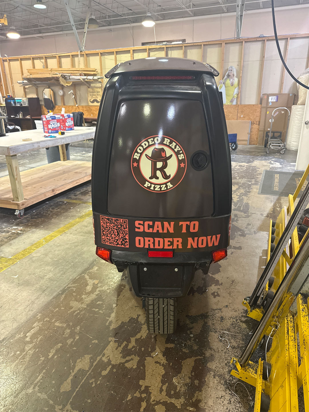 Rear view of a black delivery scooter with Rodeo Ray's Pizza logo and text 'Scan to Order Now' inside a warehouse.