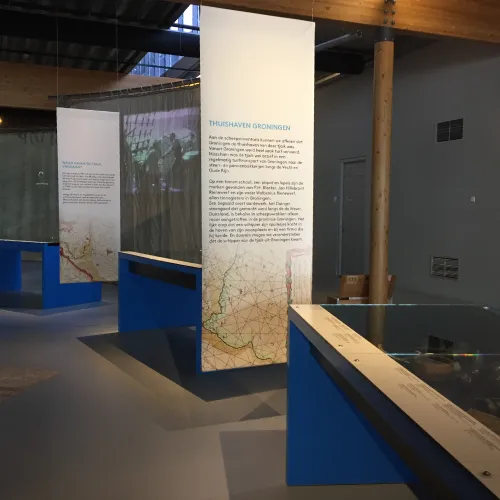 Museumtentoonstelling met informatiepanelen en kaarten die aan het plafond hangen en vitrines op blauwe standaards.