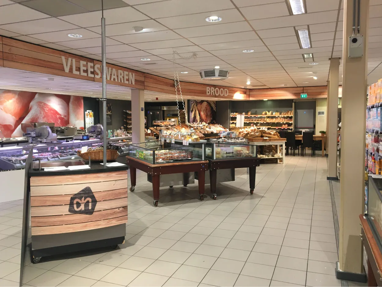 Interieur van een supermarkt met afdelingen genaamd Vleeswaren en Brood, met onder meer een vleeswarenafdeling, bakkerij-etalages en schappen.