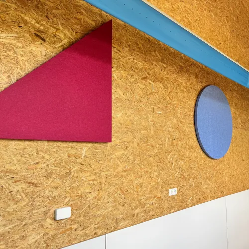 Binnenwand met structuurhouten lambrisering met een rood driehoekig akoestisch paneel en een blauw rond akoestisch paneel.