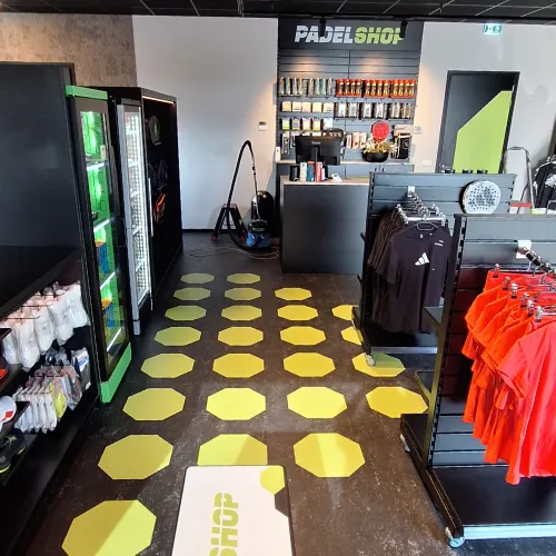 Interieur van een padelwinkel met rekken vol handschoenen, zwart-oranje shirts en een toonbank onder een bord met de tekst 'Padel Shop'.