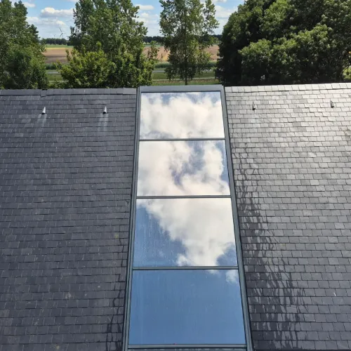 Een hoog, smal dakraam op een donker pannendak waarin de wolken en de lucht worden weerspiegeld, met bomen op de achtergrond. Beplakt met Zonwerende folie