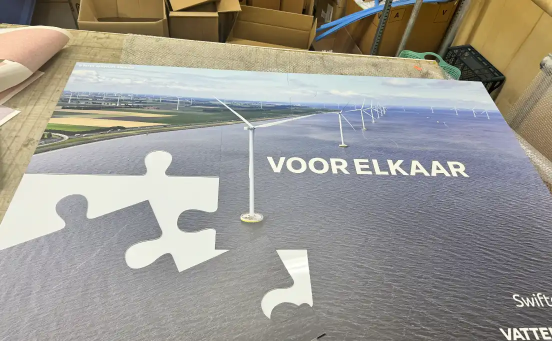 Poster van een kustwindpark met windturbines op zee en puzzelstukken over een deel van het water, met de Nederlandse tekst ‘VOOR ELKAAR’ duidelijk zichtbaar.